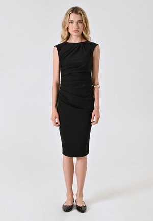 Black, sleeveless dress with gathered detailing at the waist, knee-length. Pair of black flats and a simple gold cuff bracelet worn on left arm.