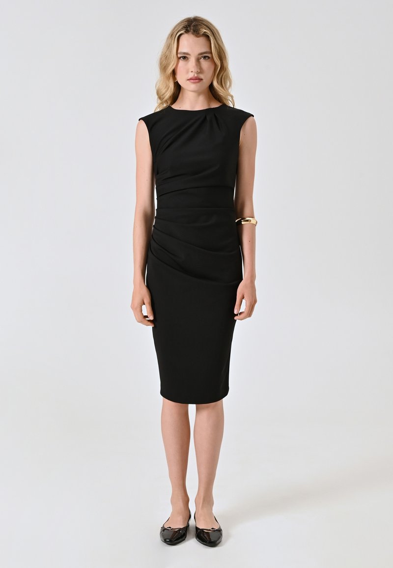 Robe noire sans manches avec des détails froncés à la taille, longueur genoux. Une paire de ballerines noires et un simple bracelet en or porté au bras gauche.