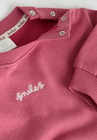 Rosa sweatshirt med ribbad krage och ärmslut, med silverknappar vid halsringningen och vit broderad text som säger "smiles."