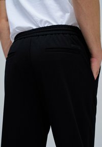 Pantalones de chándal negros con un tejido texturizado, cintura elástica y bolsillos laterales. Una pequeña etiqueta de la marca es visible cerca de la cintura.