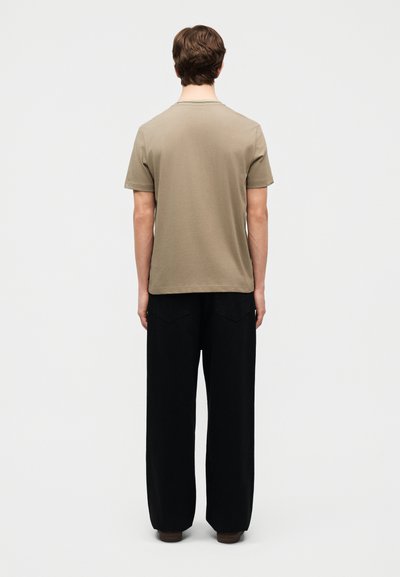 Νεαρός άνδρας που στέκεται με την πλάτη γυρισμένη, φορώντας μια απλή μπεζ κοντομάνικη μπλούζα και μαύρο παντελόνι με φαρδιές παντελόνες, σε ουδέτερο φόντο.