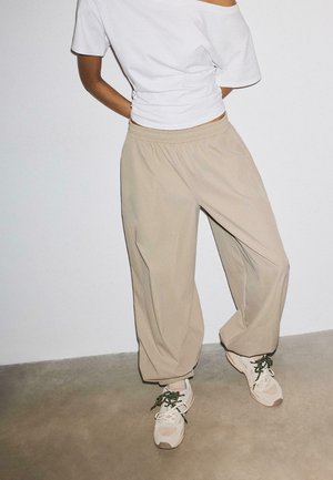 Persoon die losse beige broek met elastische manchetten, wit off-shoulder top en lichtbeige sneakers draagt, staand op een neutrale vloer tegen een witte muur.