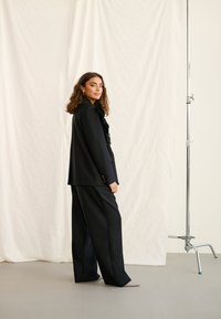 Ženska s kodrastimi lasmi, oblečena v črno obleko z nabrano bluzo, stoji bočno ob belem tekstilnem ozadju in kovinskem stojalu.