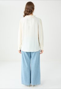 Jasny kremowy bluzka z długim rękawem i falbaniastym dekoltem, w połączeniu z szerokimi jasnoniebieskimi dżinsami. Gładki materiał i luźny krój.