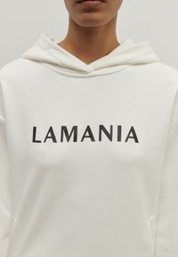 Biely mikina s kapucňou a šnúrkou, so slovom "LAMANIA" v tučných čiernych písmenách na prednej strane. Hladká textúra látky.