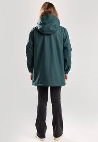 Teal waterproof jacket with a hood, elastic cuffs, and a long design, worn over dark flared jeans, viewed from the back.
