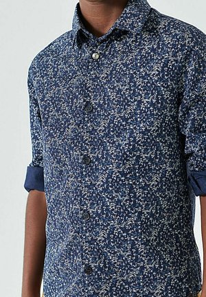 Blå skjorte med knapper, lavet af let stof med et blomstret mønster i hvidt og lyseblåt, med opkrogede ærmer og en traditionel krave.