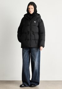 Musta puhuva takki hupulla, vetoketjukiinnityksellä, nyöri vyötäröllä ja kahdella etutaskulla. Päällä musta neule ja väljät siniset farkut.