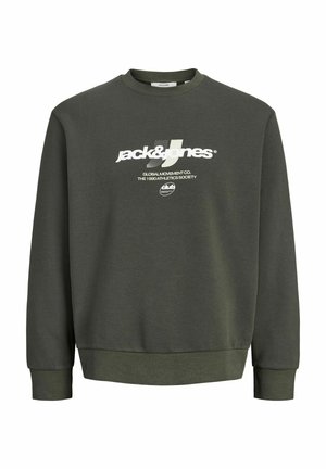 Ung person iført en mørkegrøn sweatshirt, sorte jeans, gule støvler og en sort kasket, stående mod en ensfarvet lys baggrund.