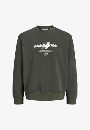 Ung person iført en mørkegrøn sweatshirt, sorte jeans, gule støvler og en sort kasket, stående mod en ensfarvet lys baggrund.