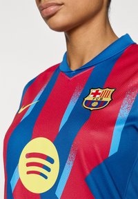 Μπλούζα της FC Barcelona με κόκκινες και μπλε κάθετες ρίγες, στρογγυλό κίτρινο λογότυπο της Spotify και έμβλημα με ασπίδα που έχει οριζόντιες ρίγες και μπάλα ποδοσφαίρου.