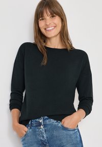 Czarny sweter dzianinowy z okrągłym dekoltem i długimi rękawami, połączony z jasnoniebieskimi dżinsami. Posiada luźny krój i prążkowane mankiety.