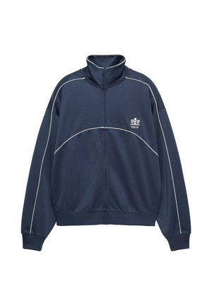 Jakna tamnoplave boje s patentnim zatvaračem i visokim ovratnikom, bijelim ukrasnim linijama i logotipom na lijevoj strani prsa. Izrađena od laganog, teksturiranog materijala.