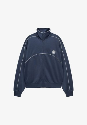 Jakna tamnoplave boje s patentnim zatvaračem i visokim ovratnikom, bijelim ukrasnim linijama i logotipom na lijevoj strani prsa. Izrađena od laganog, teksturiranog materijala.