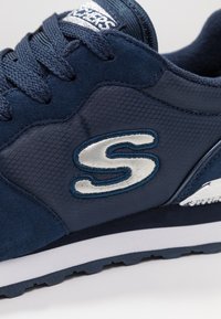 Nära bild av en mörkblå sneaker med texturerad tyg, mockaöverdrag, vit "S"-logotyp och vit mittsula på en vit yta.