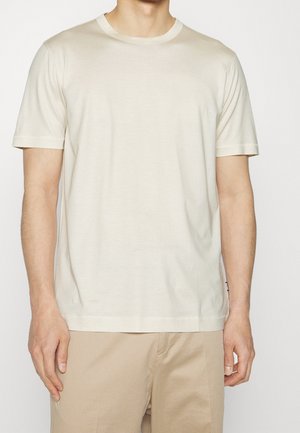 Man draagt een effen crèmekleurig T-shirt met korte mouwen en lichtbeige broek, staand tegen een neutrale achtergrond.