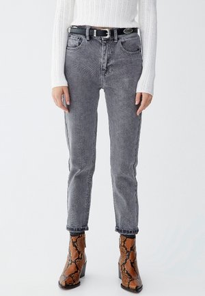 Person wearing gray high-waist jeans, black belt, white knit sweater, and brown snakeskin ankle boots standing against a plain background.
