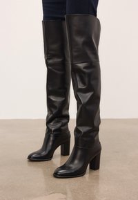 Melni ādas zābaki līdz ceļgalam ar masīvu papēdi, uzvilkti virs tumšiem biksēm, stāv uz pulēta betona grīdas.