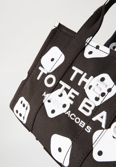 Crna platnena torba s bijelim kockastim oblicima i ugraviranim natpisima "THE TOTE BAG" i "MARC JACOBS." Čvrste ručke i strukturirani dizajn.