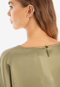 Top verde oliva con una texture liscia, dotato di un dettaglio con bottoni sul retro e un'ampia scollatura. Accessori: orecchino a clip con strass.