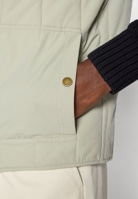 Ανοιχτό πράσινο επενδυμένο μπουφάν με χρυσή μεταλλική λεπτομέρεια λογότυπου, ριγέ μαύρα μανίκια και πλάγια τσέπη με ραφές.