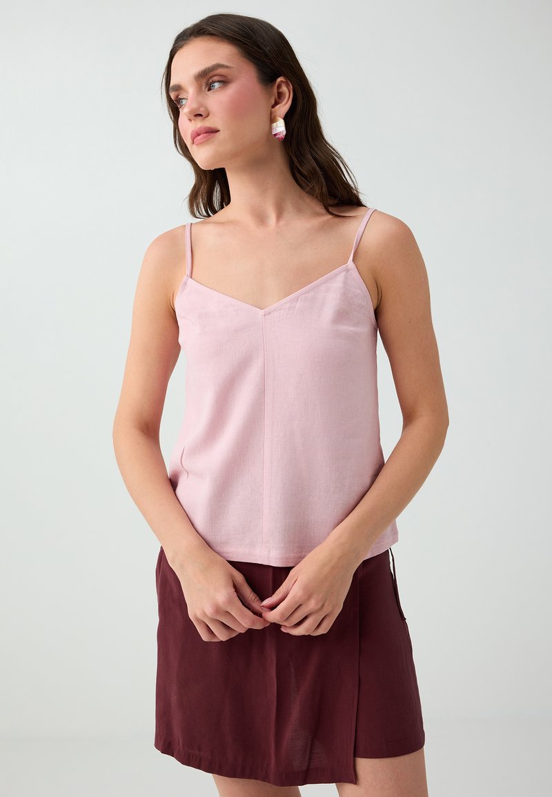 Jeune femme aux longs cheveux foncés, portant un haut rose sans manches et une jupe grenat, debout devant un fond clair uni, les mains légèrement jointes.
