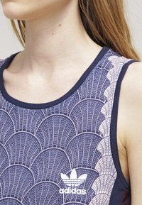 Námořnický tílko s propracovaným krémovým vroubkovaným vzorem. Má kulatý výstřih a bílou logo Adidas na spodní části. Měkká textura látky.