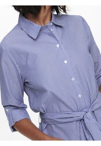Camicia a righe blu e bianche con colletto a pulsante, maniche a tre quarti e cintura legata in vita. Presenta bottoni simili a perle e tessuto morbido.