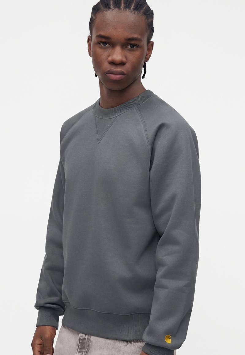 Junger Mann mit geflochtenem Haar, bekleidet mit einem schlichten dunkelgrauen Langarm-Sweatshirt und hellen Hosen, stehend vor einem weißen Hintergrund.