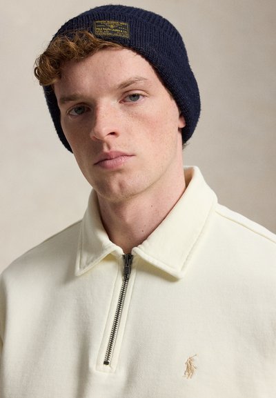 Jeune homme aux cheveux roux bouclés portant un bonnet en tricot bleu marine et un pull crème à col zippé avec un petit logo brodé sur la poitrine.