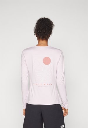 Osoba widziana od tyłu, ubrana w jasnoróżową koszulę z długim rękawem z różowym motywem słońca i fali oraz tekstem „Columbia Sportswear”.
