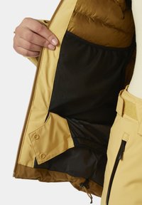 Gelbe isolierte Jacke mit einer schwarzen Netzinnentasche, die eine glatte Textur und Reißverschlüsse sowie Druckknopfakzente aufweist.