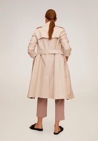 Žena s červenými vlasy v culíku, oblečená v béžovém trenčkotě, zkrácených mauvových kalhotách a černých nazouvacích sandálech, stojící na jednobarevném pozadí.