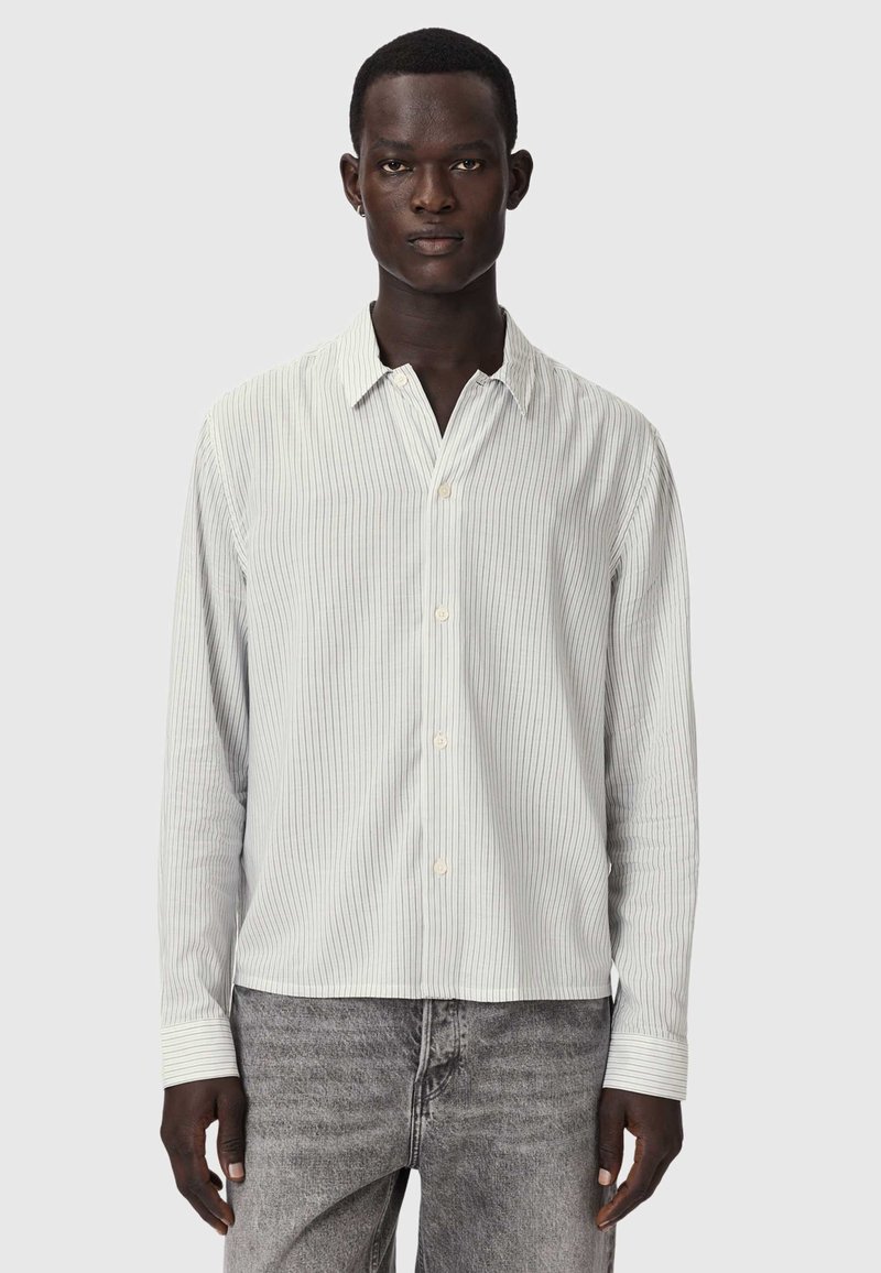 Man wearing a white long-sleeve button-up shirt with thin vertical stripes and grey washed jeans, standing against a plain background.