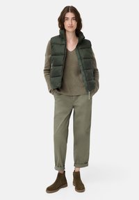 Grøn dunvest over en struktureret beige sweater, parret med rullede grønne bukser og brune ankelstøvler, mod en hvid baggrund.