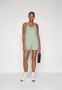 Menta zelena sportska bodysuit sa kontrastnim šavovima, racerback naramenicama i Nike logom. Model drži crnu bocu za vodu.