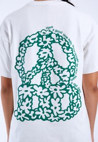 Bílé tričko s velkým zeleným symbolem míru vytvořeným z květinových tvarů. Text obklopuje design: "Užij si dobrý čas s dobrými lidmi."