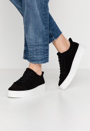 Person iført korte blå jeans og sorte platformsneakers med hvide tykke såler, den ene fod let løftet.