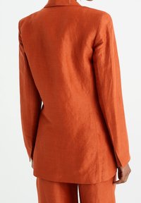 Dlouhý oranžový lněný blazer s špičatým límcem a dlouhými rukávy. Má hladkou texturu a přiléhavý střih s postranními rozparky na spodním lemu.