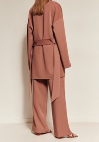 Woman standing against plain wall wearing loose brown belted jacket, matching wide-leg pants, and square-toe sandals with block heels.