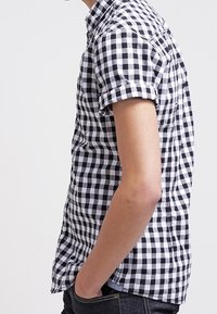 Camisa de manga corta a cuadros en blanco y negro, hecha de una tela ligera, con cuello abotonado y mangas enrolladas.