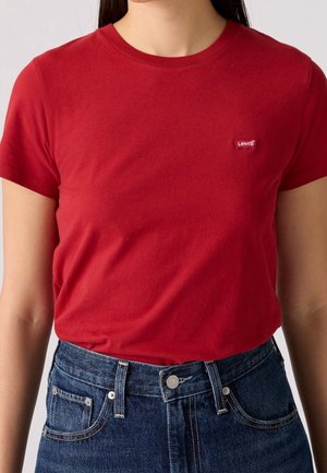 Kobieta w czerwonym t-shircie Levi's z okrągłym dekoltem, wpuszczonym do wysokich niebieskich dżinsów.