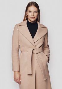 Jasnobeżowy płaszcz z paskiem, z wywijanym kołnierzem, długimi rękawami, kieszeniami bocznymi i miękką teksturą, noszony na czarnym golfie.