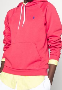 Felpa corallo con cappuccio, lacci bianchi e tasca frontale. Presenta un piccolo logo blu e polsini a coste, indossata su una maglietta gialla.
