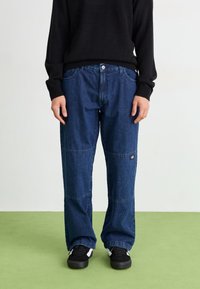 Löst sittande blå denimjeans med synlig sömnad, fem fickor och en logotyp på sidan, kombinerat med en svart tröja och sneakers.