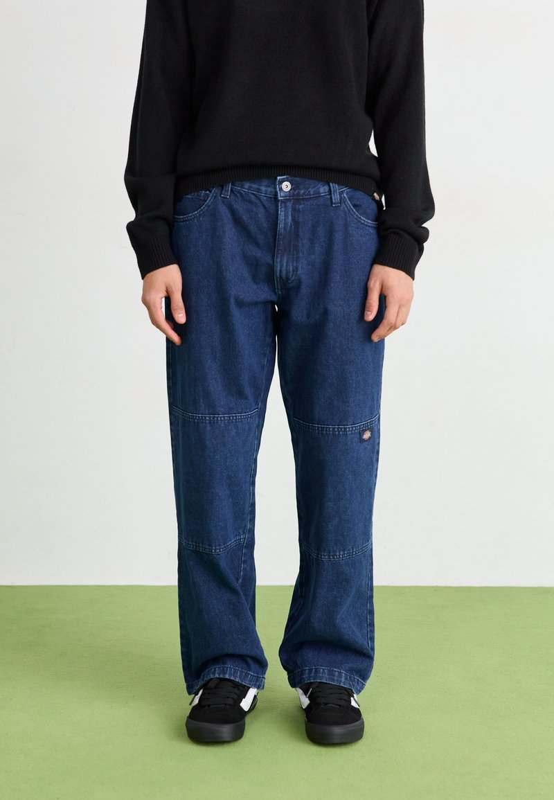 Löst sittande blå denimjeans med synlig sömnad, fem fickor och en logotyp på sidan, kombinerat med en svart tröja och sneakers.