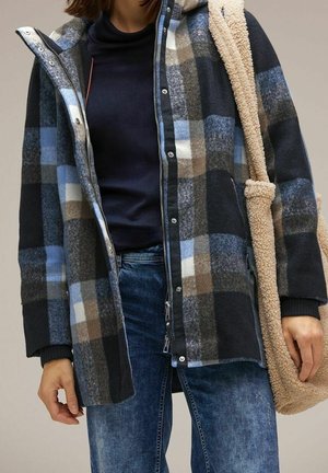 Donna che indossa una giacca a quadri blu, grigia e marrone sopra una maglietta nera, jeans blu e porta una borsa a tracolla beige e pelosa.