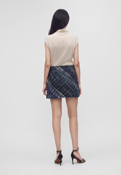 Femme aux longs cheveux foncés portant un haut beige sans manches, une mini-jupe bleue à motifs et des sandales noires à talons hauts, debout de dos.