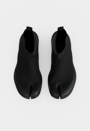 Paire de bottines noires à bout fendu avec semelles arrondies sur un fond clair uni, vue de dessus.