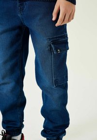 Dunkelblaue Cargo-Hose aus Denim, mit einer Seitentasche, elastischen Bündchen und einer glatten Textur, mit einem kleinen sichtbaren Markenlogo.